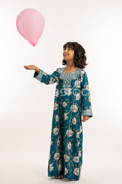 الاحتفال بالأعياد والمناسبات الدينية، فرحة وبهجة الاولاد في العيد، بورتريه لفتاة عربية خليجية اماراتية ترتدي جلابية بناتية تقوم باللعب بالبوالين، بنت سعودية تقف بتعابير السرور والسعادة، صورة لكامل الجسم، خلفية بيضاء