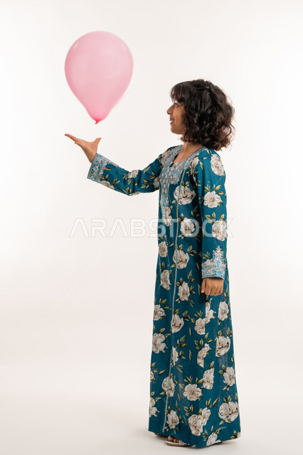 فرحة وبهجة الاولاد في العيد، الاحتفال بالأعياد والمناسبات الدينية، بورتريه لفتاة عربية خليجية اماراتية ترتدي جلابية بناتية تقوم باللعب بالبوالين، بنت سعودية انيقة تقف بتعابير السرور والسعادة، صورة لكامل الجسم، خلفية بيضاء