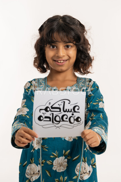 النظر الى الكاميرا بإيماءات السعادة والسرور, بورتريه مقرب لفتاة عربية خليجية اماراتية مبتسمة ترتدي جلابية بناتية تقوم بعرض ورقة بعبارة عساكم من عواده, المناسبات الدينية والأعياد الإسلامية، أجواء و طقوس العيد في الامارات, بنت سعودية انيقة تحمل لوحة، خلفية بيضاء