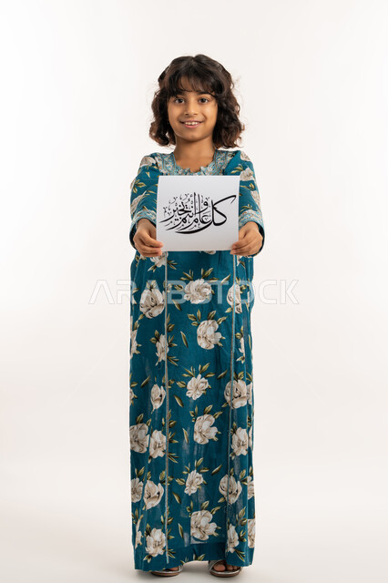 التهنئة بقدوم المناسبات الأعياد الدينية الإسلامية، إيماءات السعادة والسرور, بورتريه لفتاة عربية خليجية اماراتية مبتسمة ترتدي جلابية بناتية تقوم بعرض ورقة بعبارة كل عام وانتم بخير، بنت سعودية انيقة تحمل لوحة، صورة لكامل الجسم، خلفية بيضاء