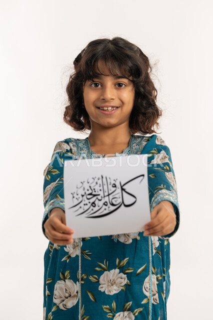 التهنئة بقدوم المناسبات الأعياد الدينية الإسلامية، النظر الى الكاميرا بإيماءات السعادة والسرور, بورتريه لفتاة عربية خليجية اماراتية مبتسمة ترتدي جلابية بناتية تقوم بعرض ورقة بعبارة كل عام وانتم بخير, بنت سعودية انيقة تحمل لوحة، خلفية بيضاء