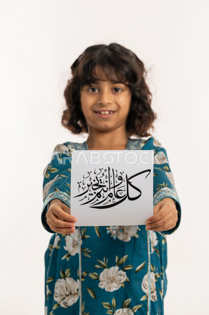 النظر الى الكاميرا بإيماءات السعادة والسرور, التهنئة بقدوم المناسبات الأعياد الدينية الإسلامية، بورتريه لفتاة عربية خليجية اماراتية مبتسمة ترتدي جلابية بناتية تقوم بعرض ورقة بعبارة كل عام وانتم بخير, بنت سعودية انيقة تحمل لوحة، خلفية بيضاء