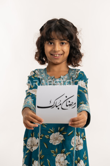 الأعياد الإسلامية والمناسبات الدينية، النظر الى الكاميرا بإيماءات السعادة والسرور, بورتريه لفتاة عربية خليجية اماراتية مبتسمة ترتدي جلابية بناتية تقوم بعرض ورقة بعبارة رمضان مبارك، التهنئة بقدوم شهر الخير, بنت سعودية انيقة تحمل لوحة، خلفية بيضاء