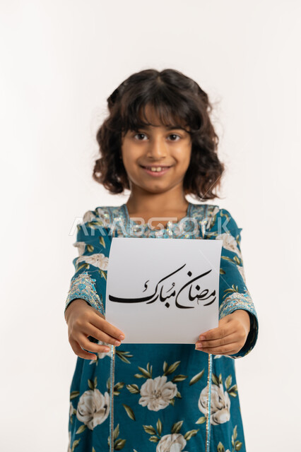 التهنئة بقدوم شهر الخير, الأعياد الإسلامية والمناسبات الدينية، النظر الى الكاميرا بإيماءات السعادة والسرور, بورتريه لفتاة عربية خليجية اماراتية مبتسمة ترتدي جلابية بناتية تقوم بعرض ورقة بعبارة رمضان مبارك، بنت سعودية انيقة تحمل لوحة، خلفية بيضاء