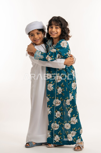 مفهوم المودة بين الاخوة, قضاء اوقات ممتعة مع العائلة، بورتريه لشقيقين عربيين خليجيين اماراتيين مبتسمان يعانقان بعضهما بإيماءات السعادة و الفرح, تقوية وتعزيز الروابط الاسرية, صورة لكامل الجسم، خلفية بيضاء