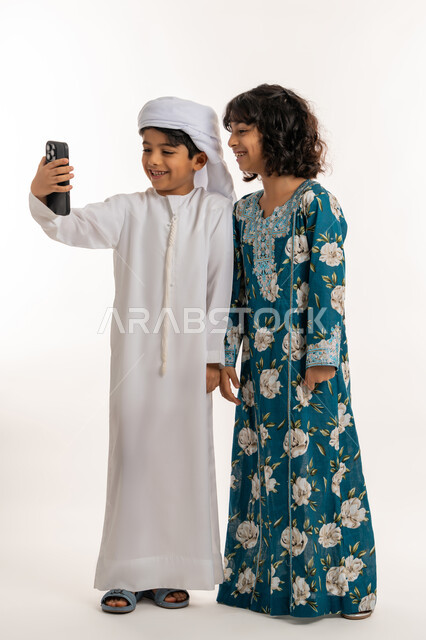 استخدام الاجهزة التقنية الحديثة، الاحتفاظ باللحظات السعيدة، ايماءات البهجة والسرور، بورتريه لولد عربي خليجي اماراتي يرتدي العمامة والكندورة يقف برفقة شقيقته يقوم بالتقاط صور السيلفي بواسطة الهاتف المحمول, صورة لكامل الجسم، خلفية بيضاء