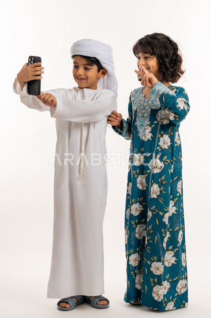 Gestures of joy and happiness, using modern technical devices, preserving happy moments, portrait of an Arab Gulf Emirati boy wearing a turban and a kandura standing with his sister taking selfies with a mobile phone, a girl raising her hand with an expression of victory, a full body photo, white background