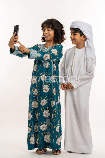 استخدام الاجهزة التقنية الحديثة، الاحتفاظ باللحظات السعيدة، ايماءات البهجة والسرور، بورتريه مقرب لبنت عربية خليجية اماراتية ترتدي جلابية بناتية تقف برفقة شقيقها تقوم بالتقاط صور السيلفي بواسطة الهاتف المحمول، صورة لكامل الجسم، خلفية بيضاء