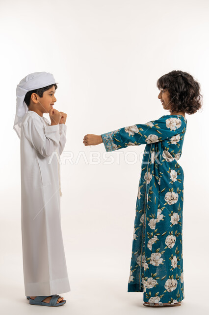 تعابير السرور والحماس، الاحتفال بقدوم العيد، ايماءات الفرحة و البهجة، المناسبات والاعياد الدينية، بورتريه من الجانب لفتاة عربية خليجية اماراتية ترتدي جلابية بناتية تقوم بتقديم العيدية لشقيقها، قضاء اوقات ممتعة مع العائلة، صورة لكامل الجسم، خلفية بيضاء