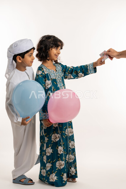 الاعياد والمناسبات الدينية، الاحتفال بقدوم العيد، تعابير السرور والحماس، ايماءات الفرحة و البهجة، بورتريه من الجانب لشقيقين عربيين خليجيين اماراتيين يحملان البوالين الملونة يقومان بتلقي العيدية من والدهما، قضاء اوقات ممتعة مع العائلة، صورة لكامل الجسم، خلفية بيضاء