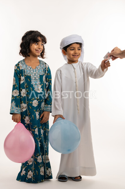 الاحتفال بقدوم العيد، تعابير السرور والحماس، ايماءات الفرحة و البهجة، المناسبات والاعياد الدينية، بورتريه لشقيقين عربيين خليجيين اماراتيين يحملان البوالين الملونة يقومان بتلقي العيدية من والدهما، قضاء اوقات ممتعة مع العائلة، صورة لكامل الجسم، خلفية بيضاء
