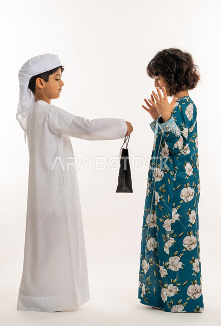 فرحة و بهجة الأطفال في العيد، تقديم الهدايا في الأعياد والمناسبات السعيدة، بورتريه من الجانب لولد عربي خليجي اماراتي يرتدي الكندورة والعمامة البيضاء يحمل بيده هدية بكيس ورقي يقدمها لبنت, ايماءات الدهشة والسرور، صورة لكامل الجسم، خلفية بيضاء