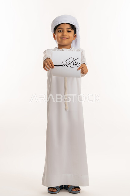 المناسبات الدينية والأعياد الإسلامية، النظر الى الكاميرا بإيماءات السعادة والسرور, بورتريه لولد عربي خليجي اماراتي مبتسم يرتدي الكندورة والعمامة يقوم بعرض ورقة بعبارة رمضان مبارك، التهنئة بقدوم شهر الخير, صورة لكامل الجسم، خلفية بيضاء