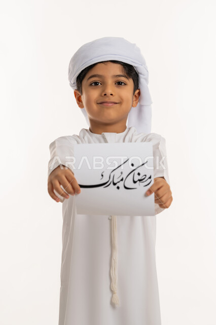 الأعياد الإسلامية والمناسبات الدينية، النظر الى الكاميرا بإيماءات السعادة والسرور, بورتريه لولد عربي خليجي اماراتي مبتسم يرتدي الكندورة والعمامة يقوم بعرض ورقة بعبارة رمضان مبارك، التهنئة بقدوم شهر الخير، خلفية بيضاء