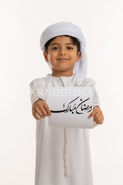 التهنئة بقدوم شهر الخير، الأعياد الإسلامية والمناسبات الدينية، النظر الى الكاميرا بإيماءات السعادة والسرور, بورتريه لولد عربي خليجي اماراتي مبتسم يرتدي الكندورة والعمامة يقوم بعرض ورقة بعبارة رمضان مبارك، خلفية بيضاء