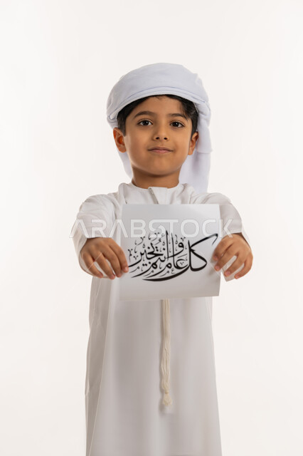 تعابير السعادة والسرور, أجواء وطقوس العيد في الامارات, المناسبات الدينية والأعياد الإسلامية، بورتريه لولد عربي خليجي اماراتي مبتسم يرتدي الكندورة والعمامة يقوم بعرض ورقة بعبارة كل عام وانتم بخير, خلفية بيضاء