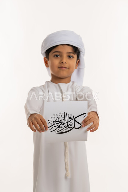 الأعياد الإسلامية والمناسبات الدينية، النظر الى الكاميرا بإيماءات السعادة والسرور, أجواء وطقوس العيد في الامارات, بورتريه لولد عربي خليجي اماراتي مبتسم يرتدي الكندورة والعمامة يقوم بعرض ورقة بعبارة كل عام وانتم بخير, خلفية بيضاء