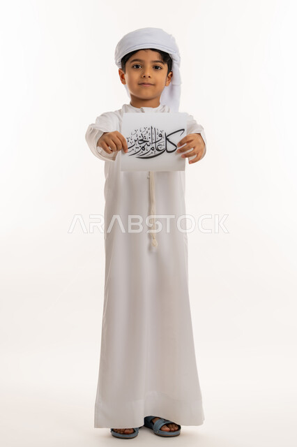 أجواء وطقوس العيد في الامارات, المناسبات الدينية والأعياد الإسلامية، النظر الى الكاميرا بإيماءات السعادة والسرور, بورتريه لولد عربي خليجي اماراتي مبتسم يرتدي الكندورة والعمامة يقوم بعرض ورقة بعبارة كل عام و انتم بخير, صورة لكامل الجسم، خلفية بيضاء