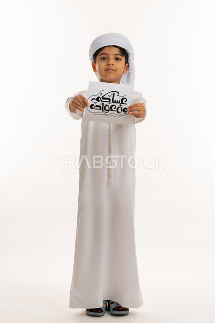 المناسبات الدينية والأعياد الإسلامية، النظر الى الكاميرا بإيماءات السعادة والسرور, بورتريه لولد عربي خليجي اماراتي مبتسم يرتدي الكندورة والعمامة يقوم بعرض ورقة بعبارة عساكم من عواده, أجواء وطقوس العيد في الامارات, صورة لكامل الجسم، خلفية بيضاء