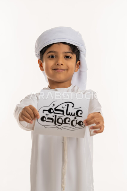 النظر إلى الكاميرا بإيماءات السعادة والسرور, بورتريه مقرب لولد عربي خليجي اماراتي مبتسم يرتدي الكندورة والعمامة يقوم بعرض ورقة بعبارة عساكم من عواده, المناسبات الدينية والأعياد الإسلامية، أجواء و طقوس العيد في الامارات, خلفية بيضاء