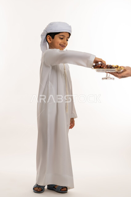 Hospitality concept, gestures of cheerfulness and welcoming guests, healthy meal of high importance, side portrait of an Arab Gulf Emirati boy wearing a kandura and turban eating dates from the host, white background