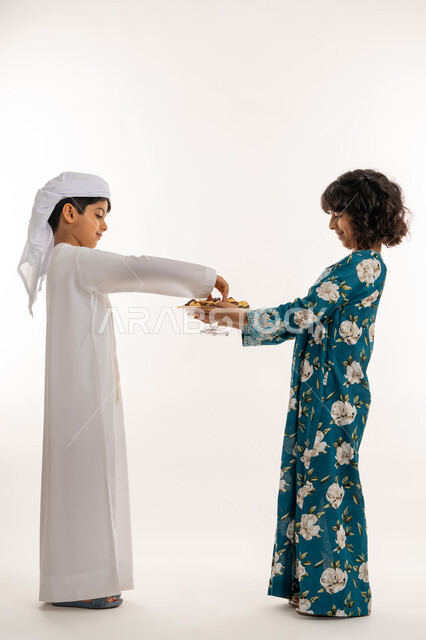ايماءات البشاشة والترحيب بالضيوف, وجبة غذائية صحية ذات أهمية عاليه, مفهوم حسن الضيافة, بورتريه من الجانب لفتاة عربية خليجية اماراتية ترتدي جلابية بناتية تقوم بتقديم التمور لولد، صورة لكامل الجسم، خلفية بيضاء