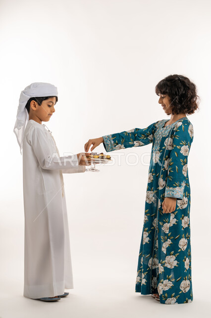 مفهوم حسن الضيافة, ايماءات البشاشة والترحيب بالضيوف, وجبة غذائية صحية ذات أهمية عاليه, بورتريه من الجانب لولد عربي خليجي اماراتي يرتدي الكندورة والعمامة بقوم بتقديم التمور لفتاة, صورة لكامل الجسم، خلفية بيضاء