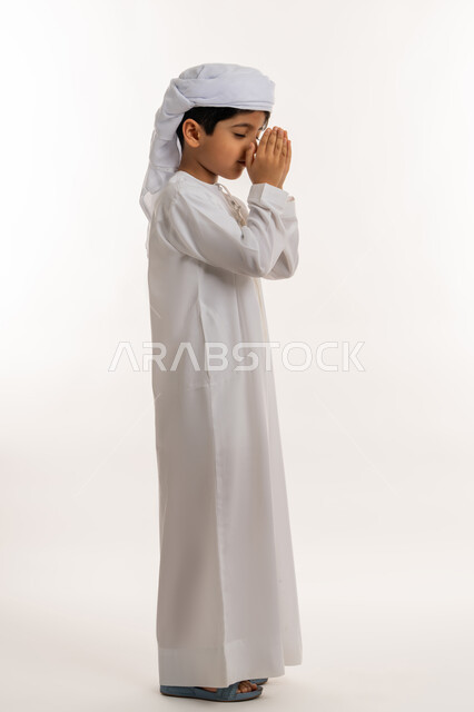 التقرب الى الله والاجتهاد في العبادة، الرغبة في تحقيق الأمنيات، بورتريه من الجانب لولد عربي خليجي اماراتي يرتدي الكندورة والعمامة يرفع يديه للاعلى بايماءات الدعاء والتضرع، صورة لكامل الجسم، خلفية بيضاء