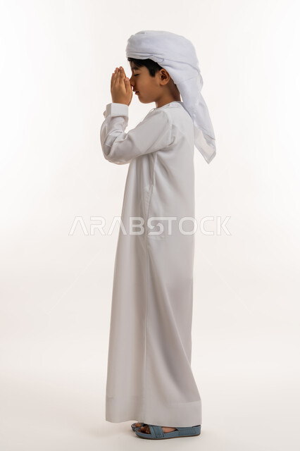 مفهوم الاجتهاد في العبادة والتقرب الى الله، الرغبة في تحقيق الأمنيات، بورتريه من الجانب لولد عربي خليجي اماراتي يرتدي الكندورة والعمامة يرفع يديه للاعلى بايماءات الدعاء والتضرع، صورة لكامل الجسم، خلفية بيضاء