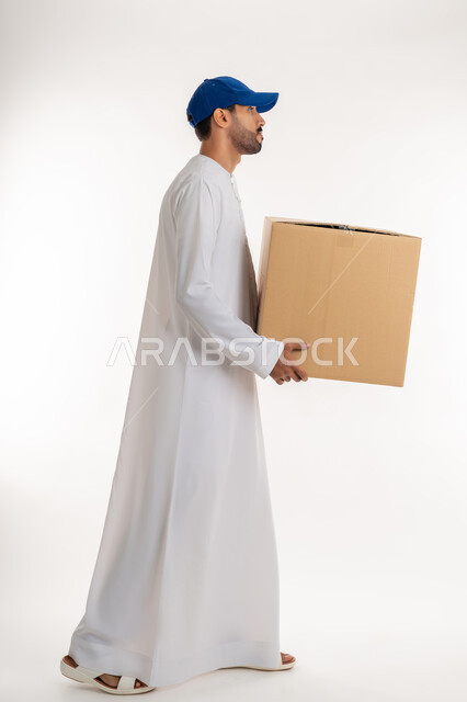 طلب  والتسوق المنتجات عبر الإنترنت، عملية توصيل الطلبات للزبائن، خدمات الشحن و التوصيل ذات الجودة العالية، بورتريه من الجانب لشاب عربي خليجي اماراتي يرتدي الكندورة و الطاقية الزرقاء يحمل صندوق كرتوني، الجسم بالطول الكامل, خلفية بيضاء