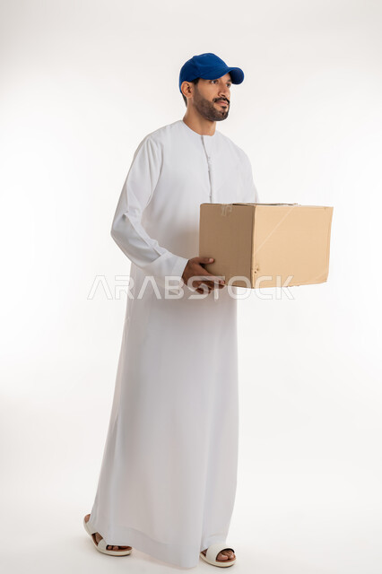 عملية توصيل الطلبات للزبائن، التسوق و طلب المنتجات عبر الإنترنت، خدمات الشحن و التوصيل ذات الجودة العالية، بورتريه من الجانب لشاب عربي خليجي اماراتي يرتدي الكندورة و الطاقية الزرقاء يحمل صندوق كرتوني، الجسم بالطول الكامل, خلفية بيضاء