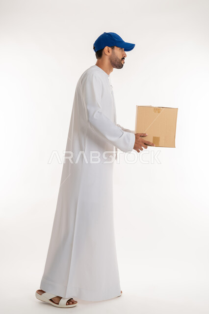 التسوق و طلب المنتجات عبر الإنترنت، عملية توصيل الطلبات للزبائن، خدمات الشحن و التوصيل ذات الجودة العالية، بورتريه من الجانب لشاب عربي خليجي اماراتي يرتدي الكندورة و الطاقية الزرقاء يحمل صندوق كرتوني، الجسم بالطول الكامل, خلفية بيضاء