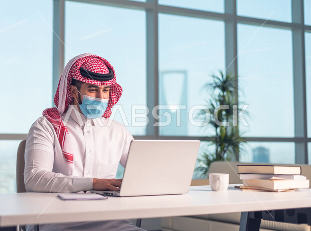 A Saudi Arabian Gulf employee working at the company’s headquarters, adhering to the precautionary precautions by wearing a mask, following up and discussing business through the laptop, achieving the company’s goals, developing work