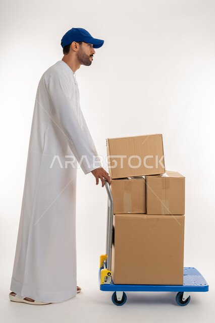 التسوق و طلب المنتجات عبر الإنترنت، إستلام المشتريات و تسليم البضائع للزبائن بالوقت المحدد و بشكل سليم, بورتريه من الجانب لمنوب توصيل عربي خليجي اماراتي يرتدي الكندورة و الطاقية الزرقاء يقوم بجر عربة مليئة بالصناديق الكرتونية، الجسم بالطول الكامل, خلفية بيضاء