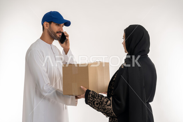 Online shopping and shopping malls, integrating advanced modern technologies into work, portrait of a young Arab Gulf Emirati man wearing casual clothes and a blue hat delivering orders standing next to an Arab Gulf Emirati woman, making a phone call to confirm reservations and delivery times, white background