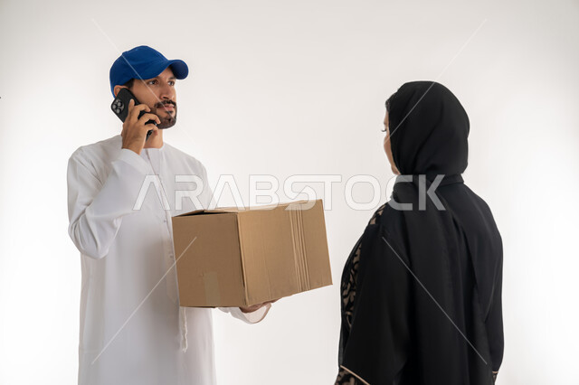 دمج التقنيات الحديثة المتطورة بالعمل، مواقع ومراكز التسوق والشراء الاونلاين, بورتريه لشاب عربي خليجي اماراتي يرتدي الزي الكاجوال و القبعة يقوم بتسليم الطلبات يقف بجانب امرأة عربية خليجية امارتيه، اجراء مكالمه هاتفيه لتأكيد الحجوزات ومواعيد الاستلام, خلفيه بيضاء