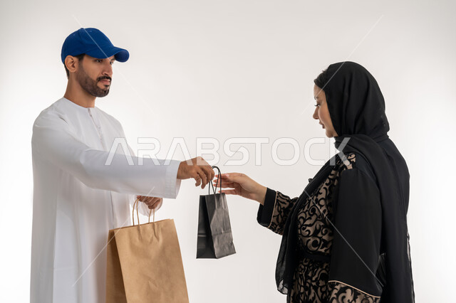التسوق وطلب المنتجات عبر الإنترنت, شبكات التوصيل الاونلاين في الخليج، دمج العمل مع التقنية والتكنولوجيا، بورتريه لشاب عربي خليجي اماراتي يرتدي الزي الكاجوال و القبعة الزرقاء يقف بجانب امرأة عربية خليجية امارتيه يقوم بتوصيل الطلبات، خلفيه بيضاء