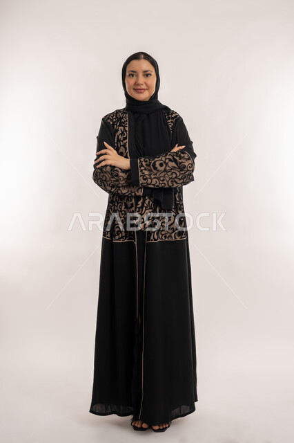 النظر الى الكاميرا بإيماءات السرور والفرح, متابعة الأزياء وصيحات الموضة, بورتريه لامرأة عربية خليجية اماراتية ترتدي العباءة السوداء تقف بأيدي مكتوفة باستقامة واستعداد, إيماءات تدل على الأنوثة والجمال, صورة الجسم بالطول الكامل, خلفية بيضاء