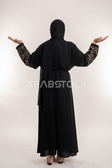 موضوع مبهم و مجهول، الاهتمام بالمظهر الخارجي, بورتريه من الخلف لامرأة عربية خليجية اماراتية محجبة ترتدي العباءة السوداء ترفع يديها بكفوف مفتوحة نحو الأعلى للسؤال عن أمر ما، صورة الجسم بالطول الكامل, خلفية بيضاء