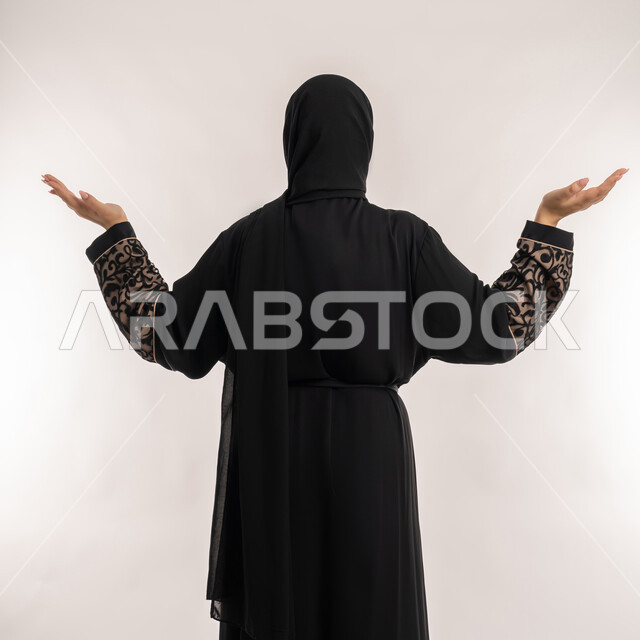 الاهتمام بالمظهر الخارجي, بورتريه من الخلف لامرأة عربية خليجية اماراتية محجبة ترتدي العباءة السوداء ترفع يديها بكفوف مفتوحة نحو الأعلى للسؤال عن أمر ما، موضوع مبهم و مجهول، خلفية بيضاء