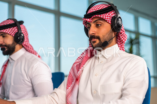 موظفان عربيان سعوديان خليجيان يعملان في خدمة العملاء في شركه يرتديان سماعة الاتصال, استعلامات واستفسارات, الاجابه على اسئلة العملاء, اجراء اتصال فيديو من خلال الحاسوب , عقد اجتماع عمل عبر الانترنت, العمل عن بعد , متابعة وتطوير العمل .