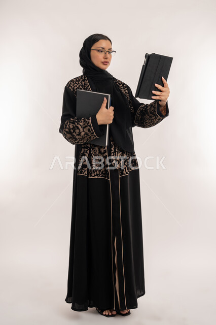 Expressions of integration and concentration, completing work tasks via tablet, portrait of a veiled Arab Gulf Emirati woman wearing a black abaya holding a notebook, using modern technical devices, women's professions and jobs, full body, white background