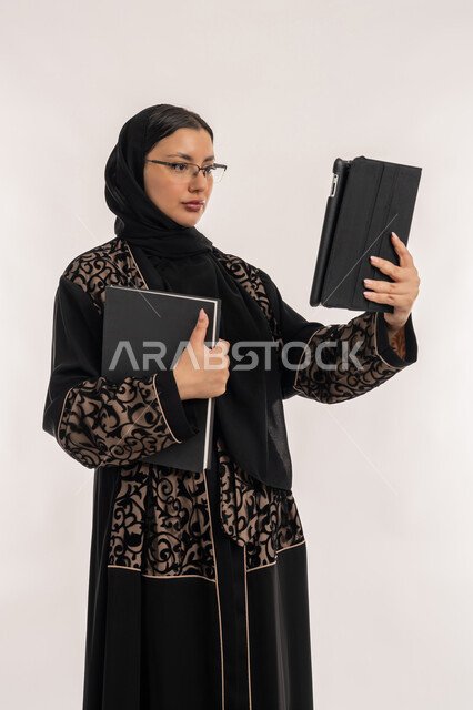 استخدام الاجهزة التقنية الحديثة, مهن ووظائف نسائية, تعابير الاندماج والتركيز، انجاز مهام العمل عبر التابليت اللوحي، بورتريه لامرأة عربية خليجية اماراتية محجبة ترتدي العباءة السوداء تحمل بيدها دفتر تدوين الملاحظات, خلفية بيضاء
