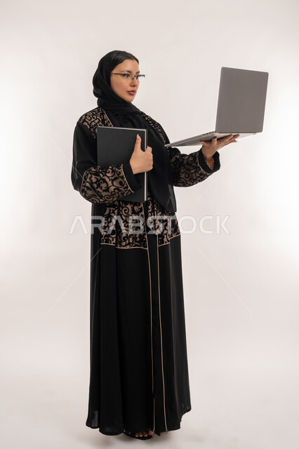 انجاز مهام العمل عبر الحاسب المحمول، تعابير الاندماج والتركيز، بورتريه لامرأة عربية خليجية اماراتية محجبة ترتدي العباءة السوداء تحمل بيدها دفتر تدوين الملاحظات, استخدام الاجهزة التقنية الحديثة, مهن ووظائف نسائية, الجسم بالطول الكامل, خلفية بيضاء
