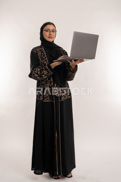 مهن ووظائف نسائية, النظر الى الكاميرا بايماءات الجدية والحزم والتركيز، بورتريه لامرأة عربية خليجية اماراتية محجبة ترتدي العباءة السوداء تقوم بانجاز مهام العمل عبر الحاسب المحمول، استخدام الاجهزة التقنية الحديثة, صورة الجسم بالطول الكامل, خلفية بيضاء