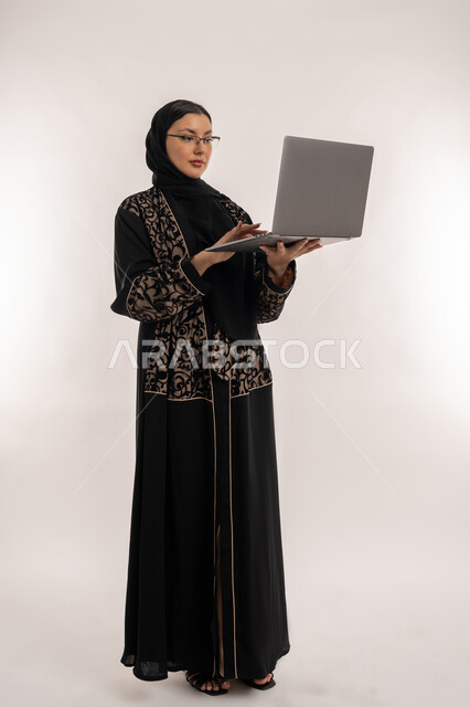 تعابير الاندماج والتركيز، مفهوم إدارة المشاريع عن بعد، بورتريه لامرأة عربية خليجية اماراتية محجبة ترتدي العباءة السوداء تقوم بانجاز مهام العمل عبر الحاسب المحمول، استخدام الاجهزة التقنية الحديثة, صورة الجسم بالطول الكامل, خلفية بيضاء