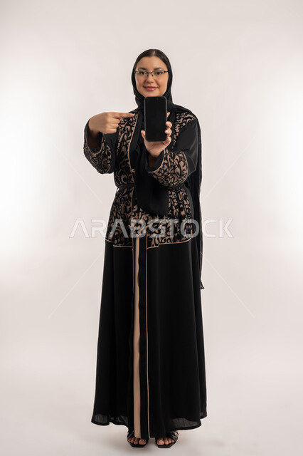 استخدام الأجهزة والتقنيات الحديثة والمتطورة، النظر الى الكاميرا بايماءات السعادة والسرور، بورتريه لإمرأة عربية خليجية اماراتية محجبة ترتدي العباءة السوداء تشير بأصبع السبابة و تقوم بعرض شاشة سوداء فارغة عبر الهاتف المحمول، صورة الجسم بالطول الكامل، خلفية بيضاء