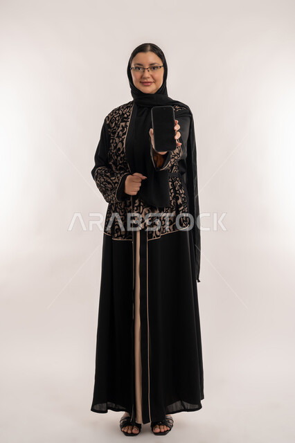 النظر الى الكاميرا بايماءات السعادة والسرور، بورتريه لإمرأة عربية خليجية اماراتية محجبة ترتدي العباءة السوداء تقوم بعرض شاشة سوداء فارغة عبر الهاتف المحمول، استخدام الأجهزة والتقنيات الحديثة والمتطورة، صورة الجسم بالطول الكامل، خلفية بيج