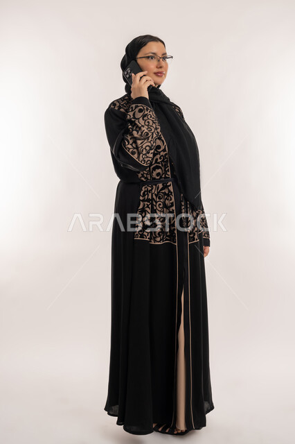 محادثات ودردشات مع الاصدقاء، تعابير الاندماج والتركيز، قضاء اوقات ممتعة بتبادل الاخبار والأحاديث، بورتريه لامرأة عربية خليجية اماراتية محجبة ترتدي العباءة السوداء تقوم بإجرء مكالمة هاتفية، الاهتمام بالمظهر الخارجي, صورة الجسم بالطول الكامل، خلفية بيضاء