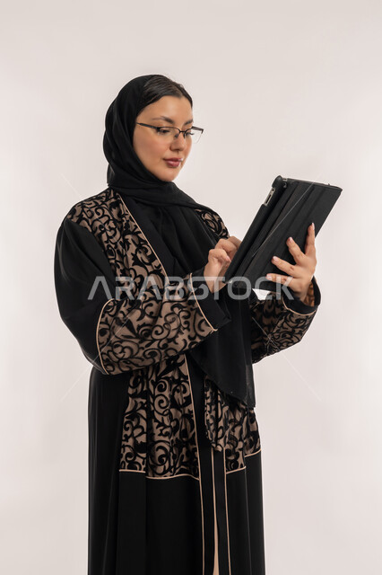 مفهوم إدارة المشاريع عن بعد، بورتريه لامرأة عربية خليجية اماراتية محجبة ترتدي العباءة السوداء تقوم بانجاز مهام العمل عبر جهاز التابلت، استخدام الاجهزة التقنية الحديثة, تعابير الاندماج والتركيز، خلفية بيضاء